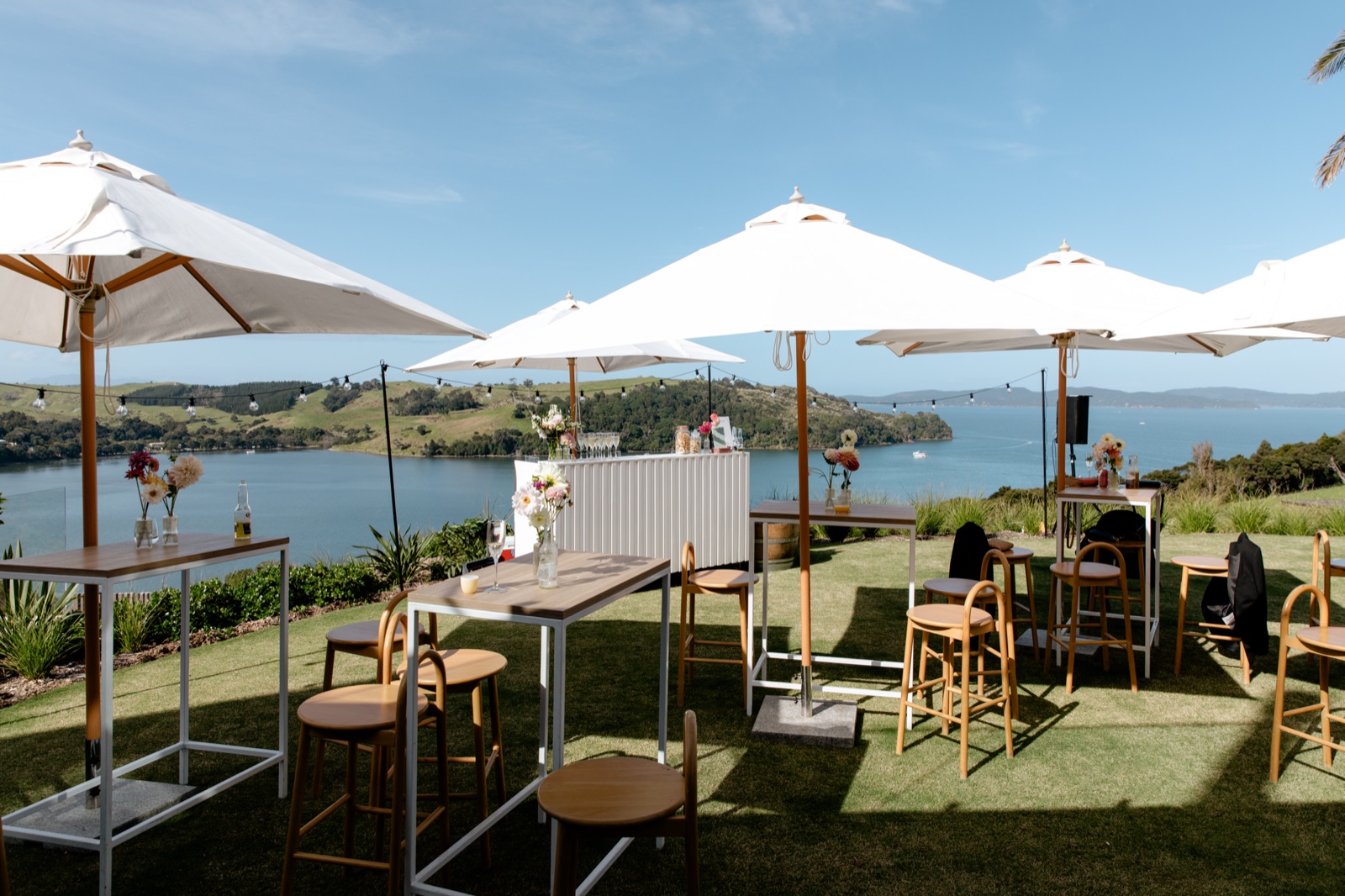 The wedding bar on the lawn at Bluestone Ridge: high tables and stools under white umbrellas, festoon lights, dahlias in bottles, and the Hauraki Gulf behind.