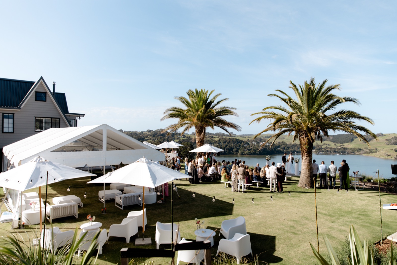 A wedding ceremony on the lawn at the Farmhouse, Bluestone Ridge: guests on white benches facing the couple at the deck, palms, a marquee with lounge seating and umbrellas, and the bay beyond.