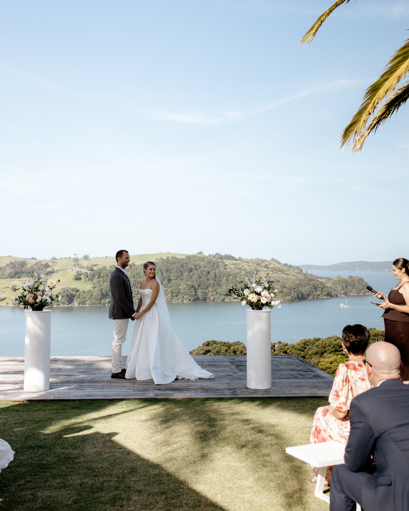 A wedding ceremony at Bluestone Ridge: the couple hand in hand on the deck above the Hauraki Gulf, the celebrant beside them, guests on white benches under the palms, green headlands and boats on the water behind.