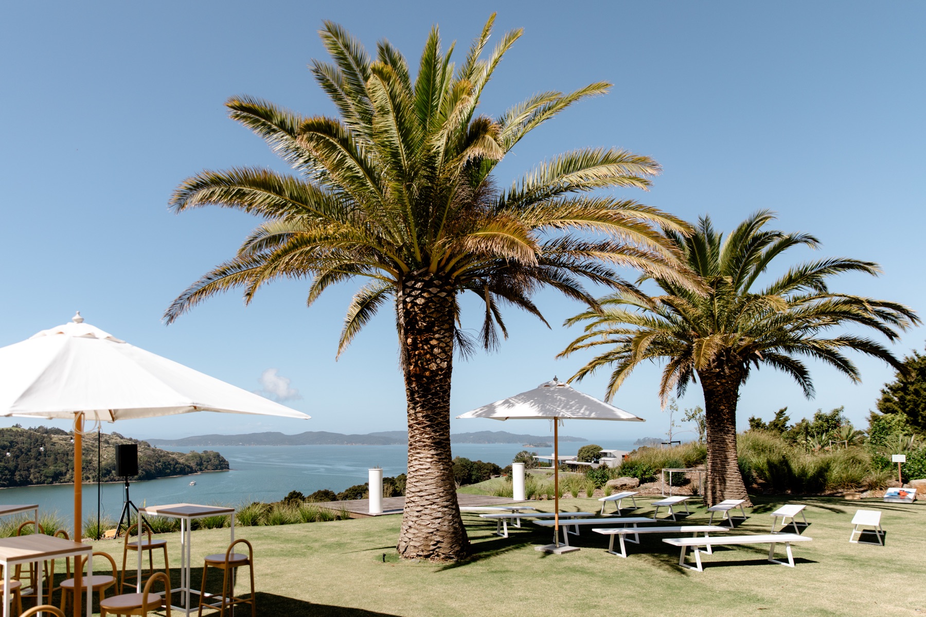 The lawn at Bluestone Ridge under two tall palms, set for a wedding with benches, umbrellas, a bar table, and the Gulf and islands behind.