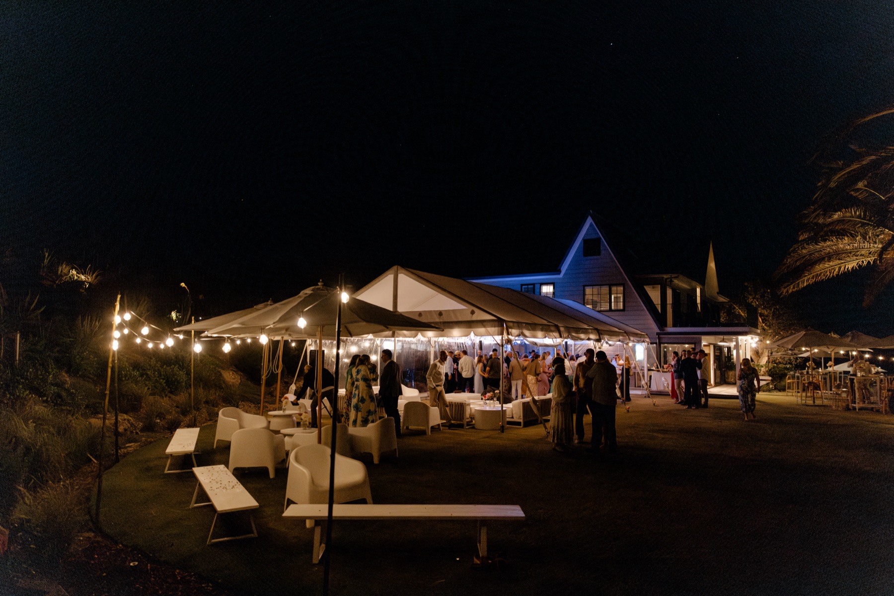 The wedding party at night at Bluestone Ridge: guests under the lit marquee and umbrellas, festoon lights along the lawn, the Farmhouse glowing behind.