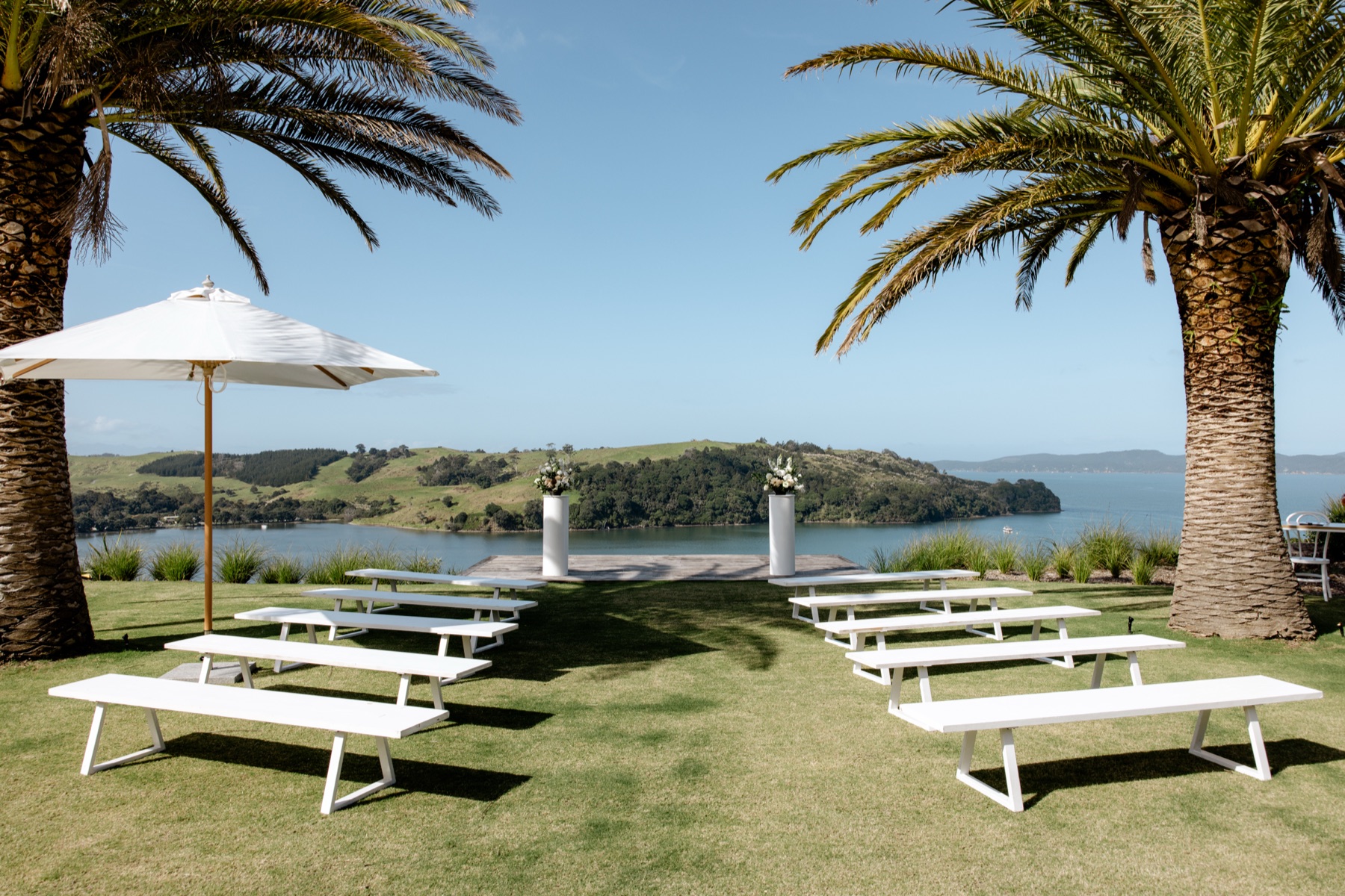 Rows of white benches on the lawn at Bluestone Ridge facing the ceremony deck, flowers on two white plinths, palms either side, and the bay and headlands beyond.
